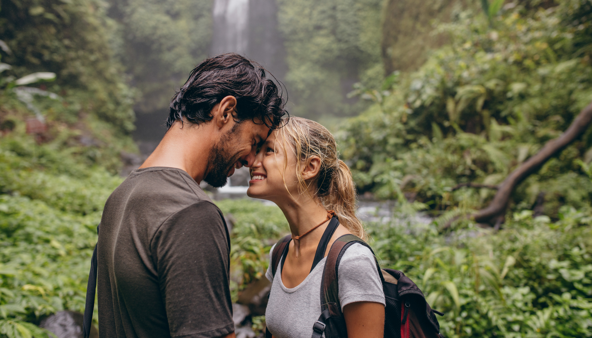 Couple touches foreheads, smiling, near a waterfall in a lush, green forest.
