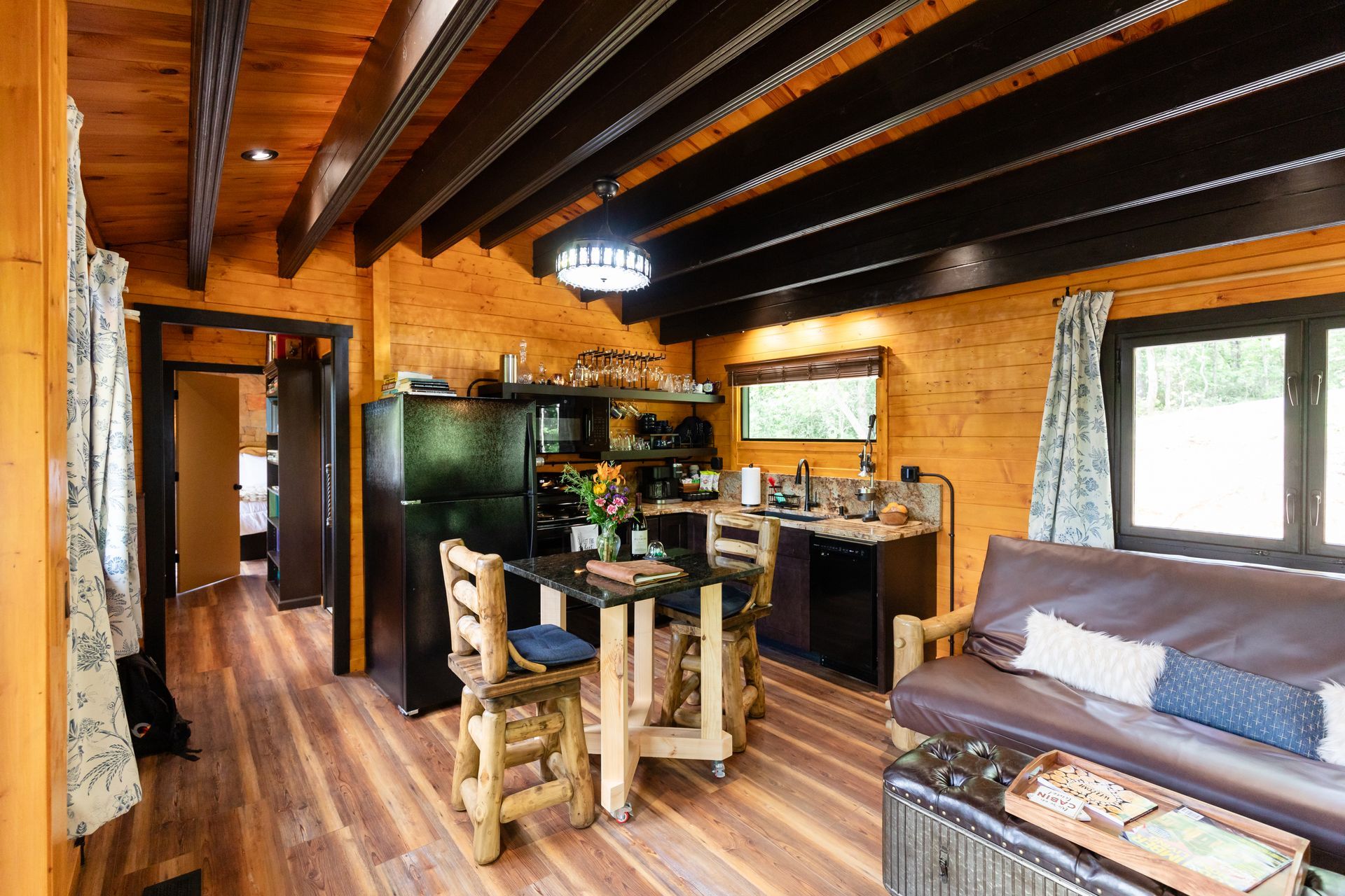 Cozy rustic cabin interior with wooden beams, kitchen, dining area, sofa, and large windows