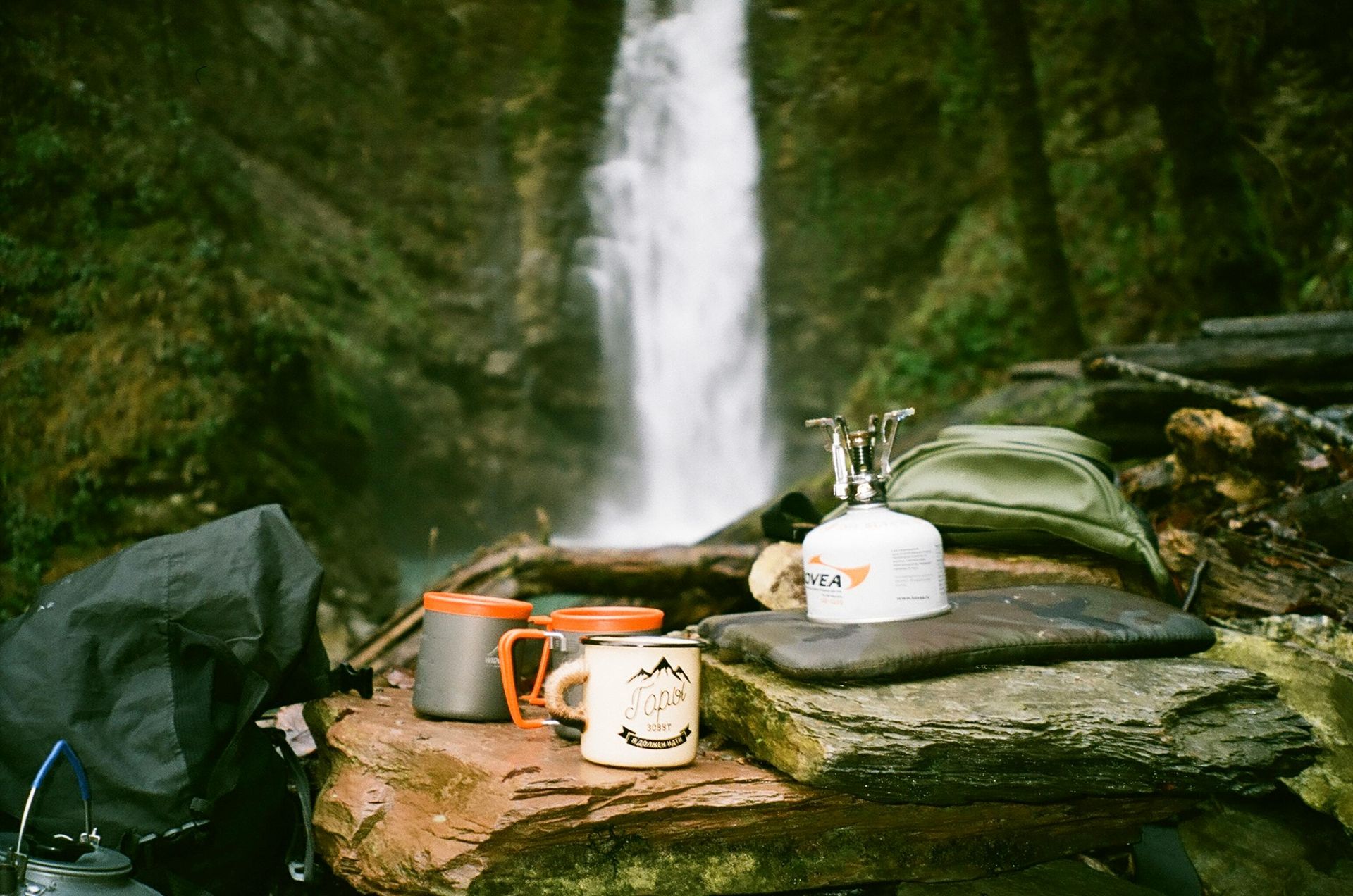 Camping setup with waterfall backdrop: mugs, camp stove, backpack, resting on rocks.