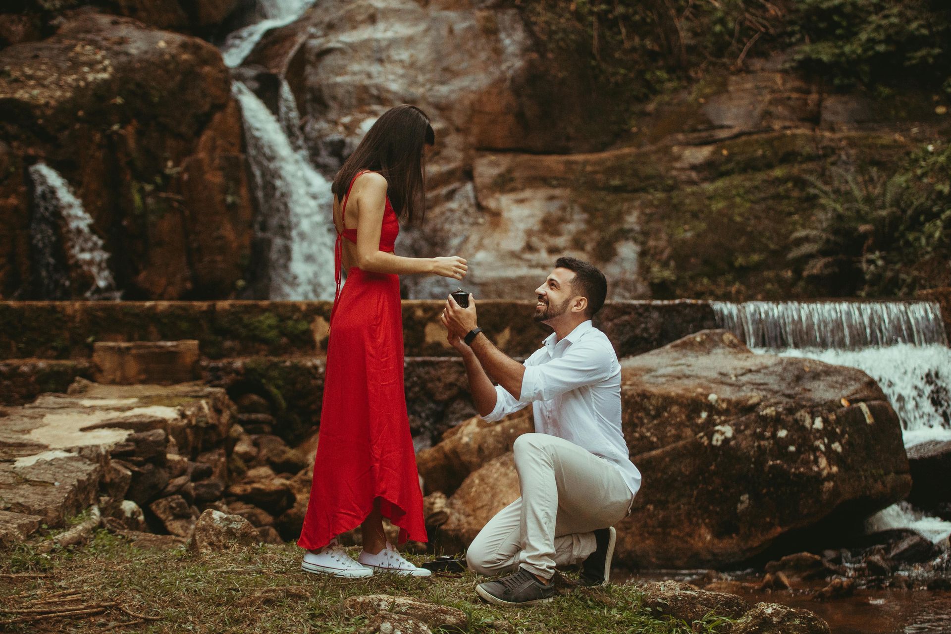 Man kneeling, proposing to a person in a red dress near a waterfall, holding a ring.