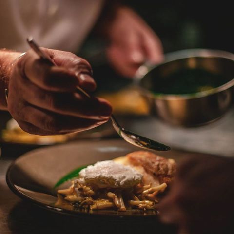 Chef garnishing a plated dish with sauce using a spoon, indoors, close-up.