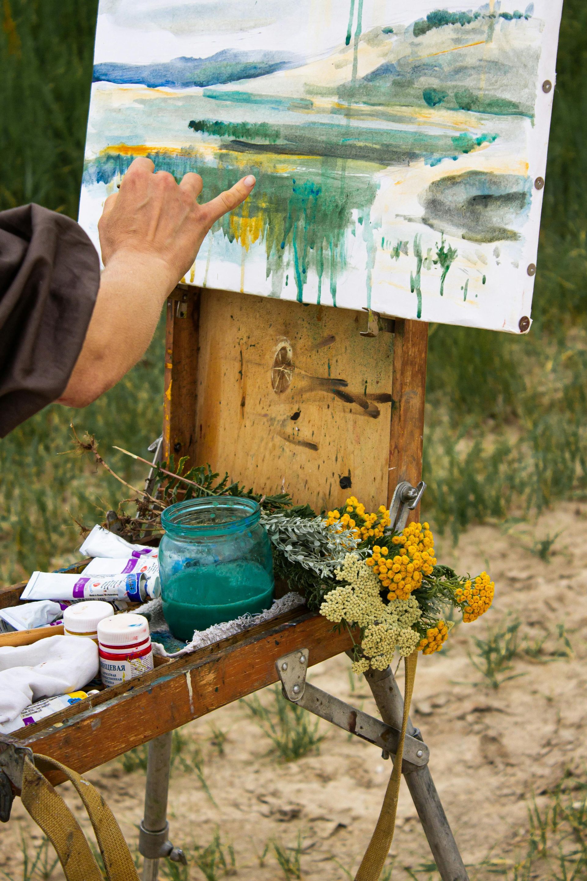 Artist painting a landscape outdoors; canvas on easel, palette, paint, flowers, hand pointing.