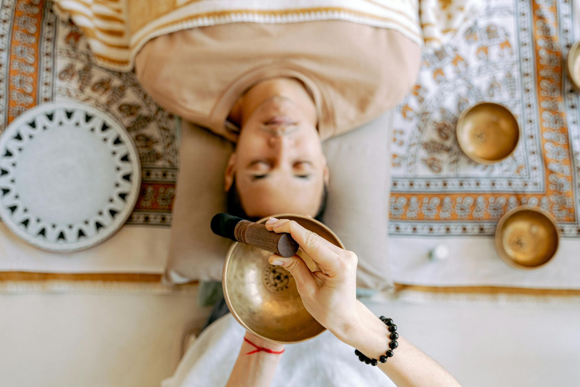 Person lying down with eyes closed, sound bowl held near head. Other bowls and decorative rug in view.