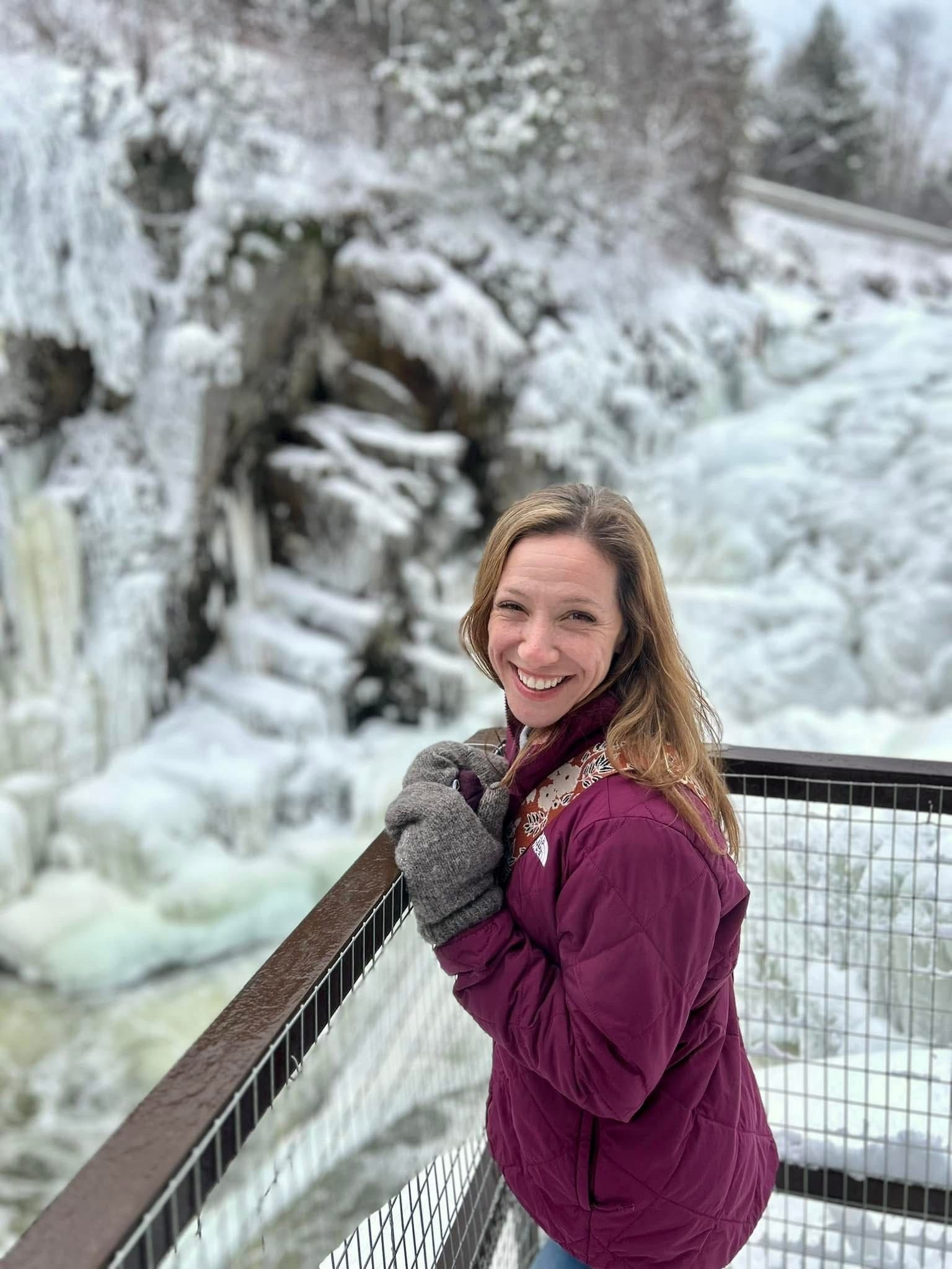 Woman in a maroon jacket smiles on a snowy bridge beside an icy waterfall.