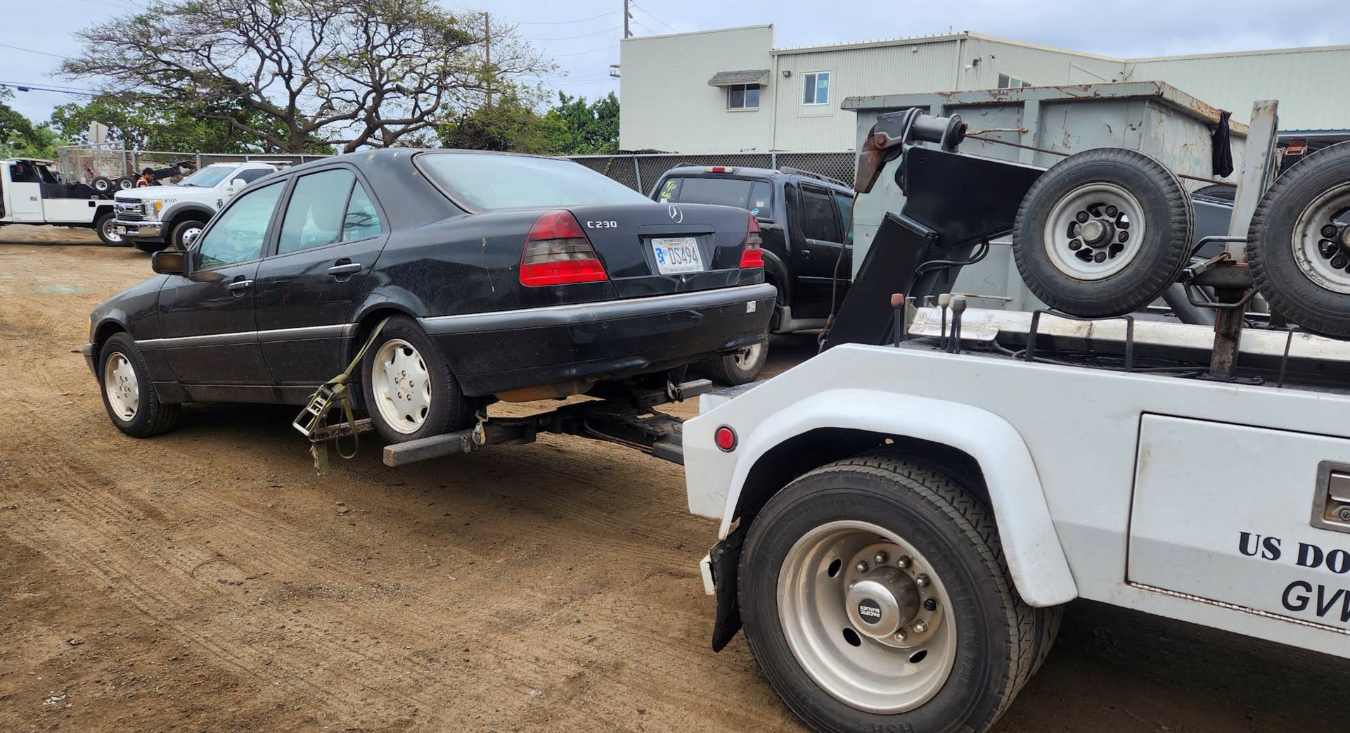 Tow truck hauling a black sedan with a damaged rear wheel in a muddy lot