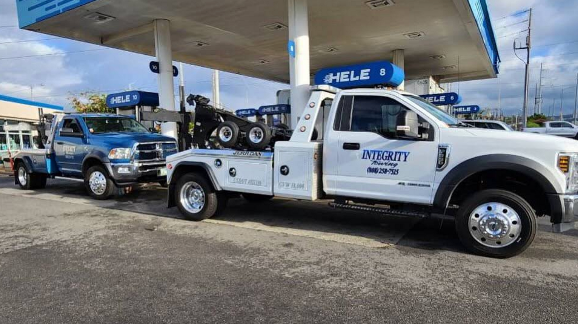 Tow trucks parked at a gas station, with one white flatbed truck in the foreground under the canopy.