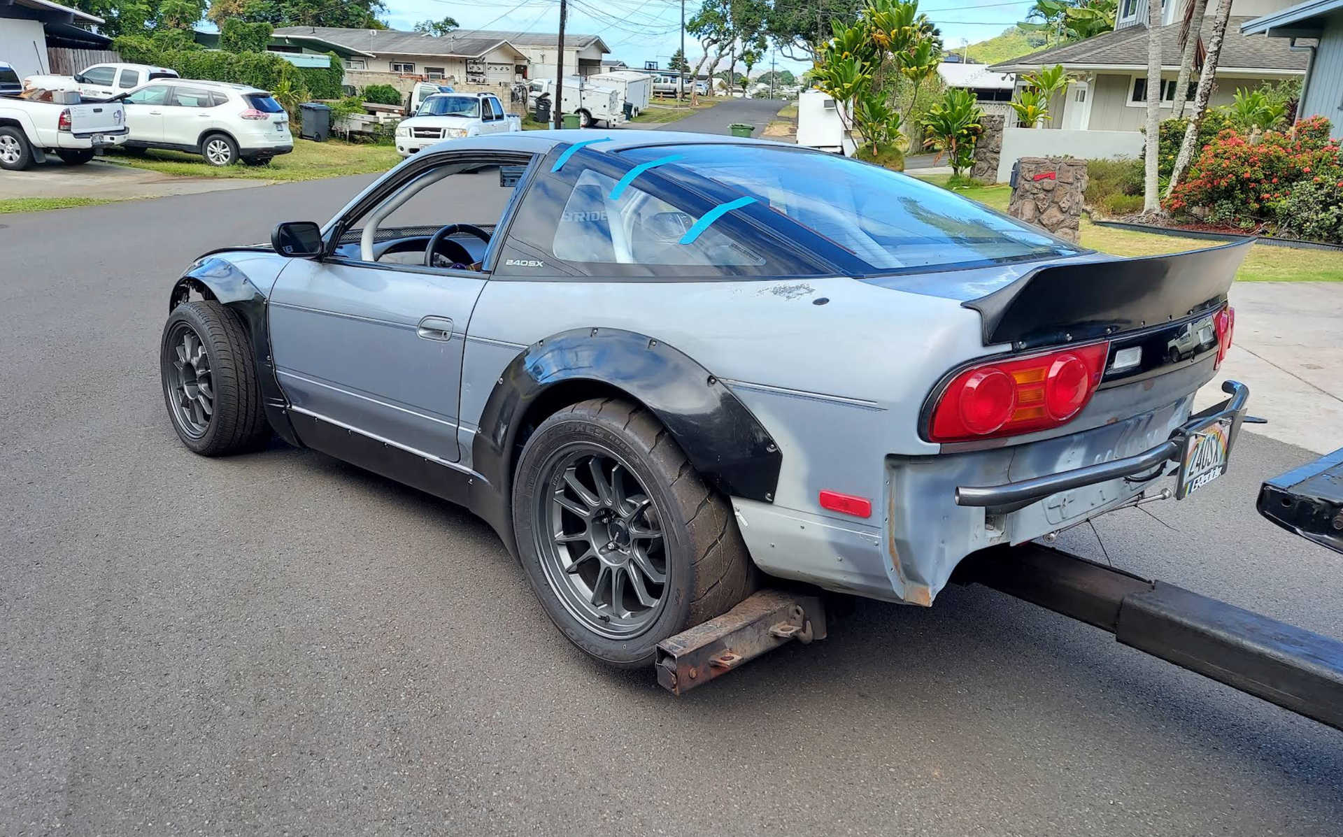 Silver sports car on a tow dolly in a driveway, rear bumper missing and driver’s side windows taped over