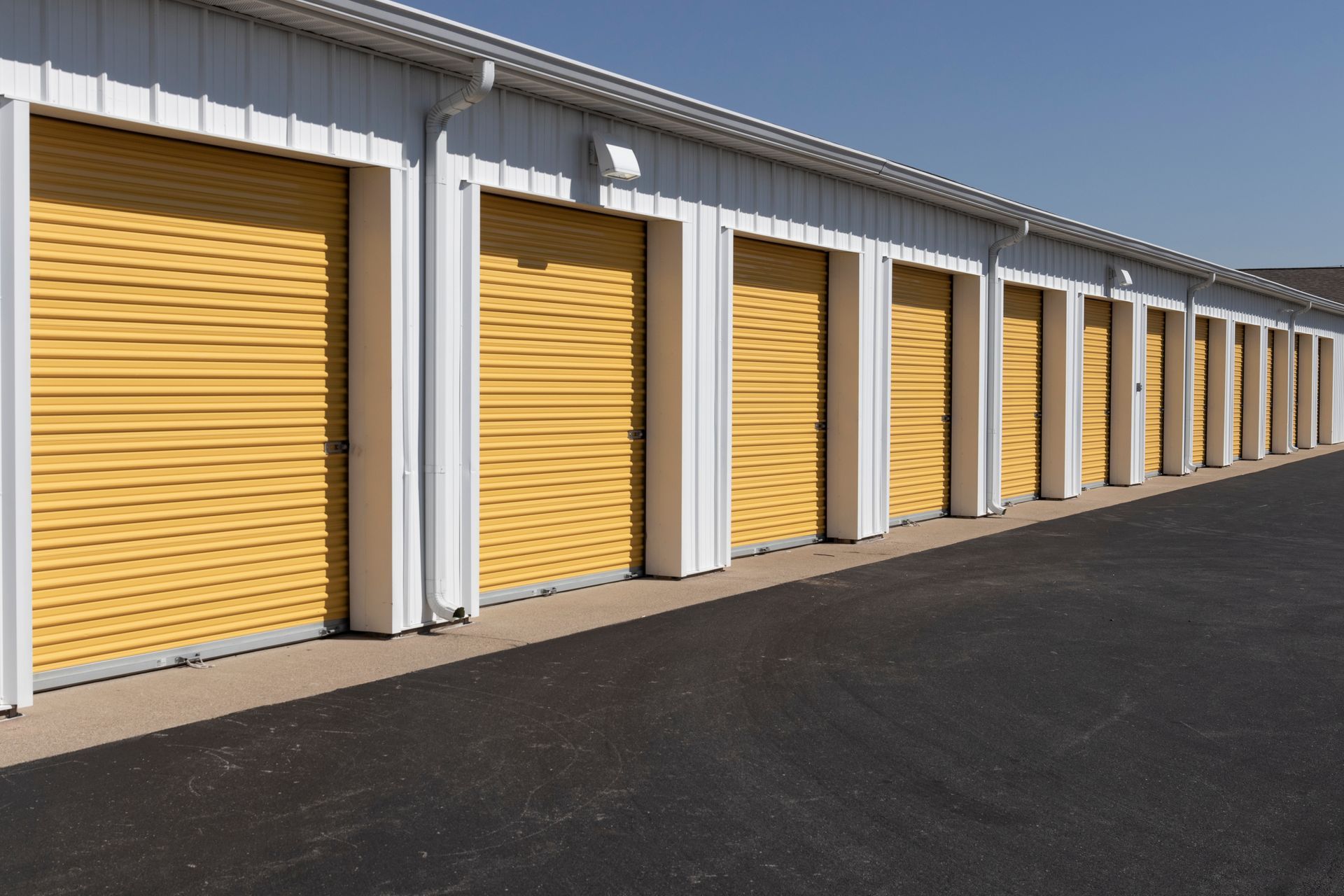 Row of yellow storage unit doors in a white building, on black asphalt, against a blue sky.