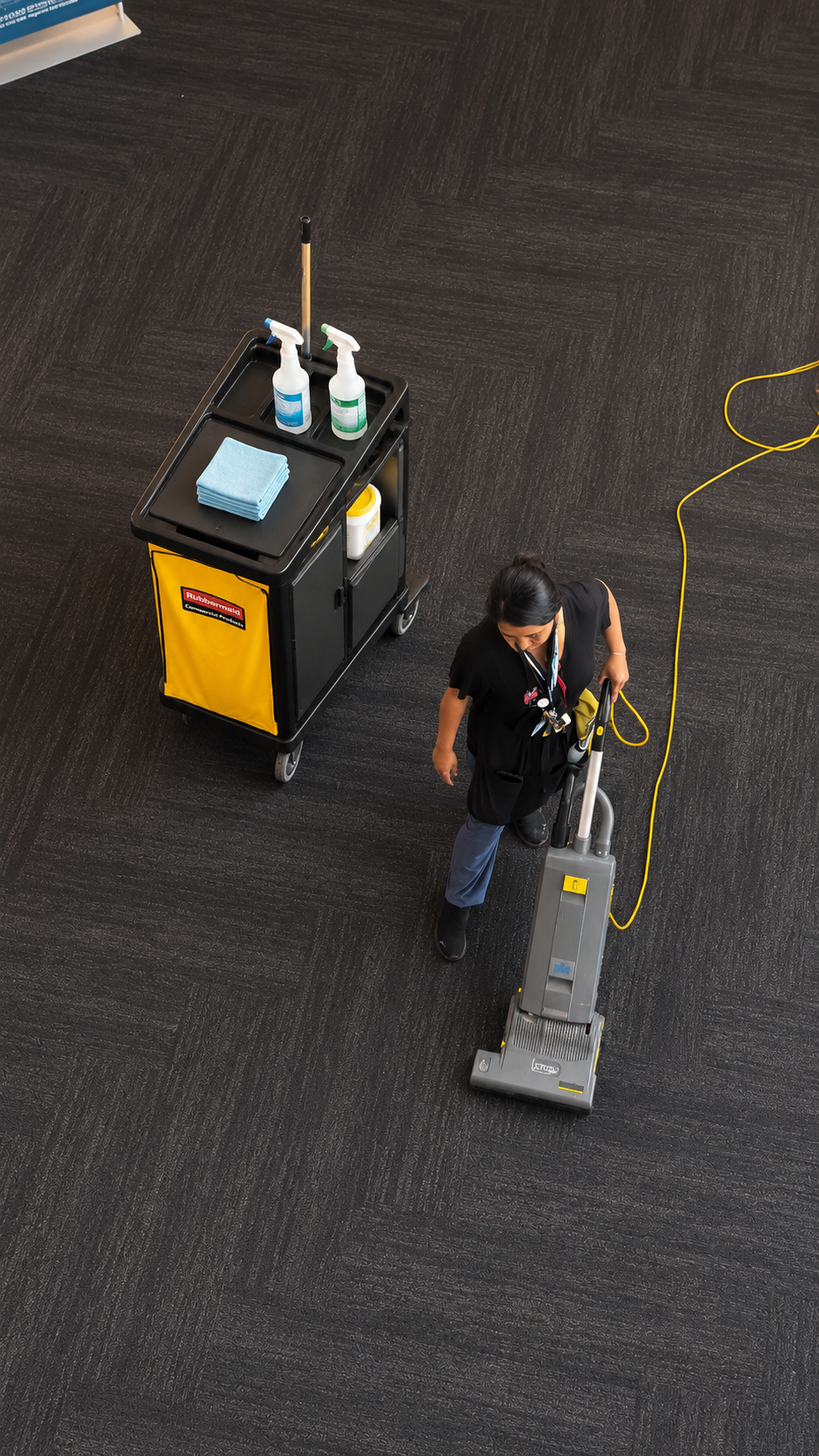 Worker operating a floor-cleaning machine on a dark carpet beside a yellow utility cart.