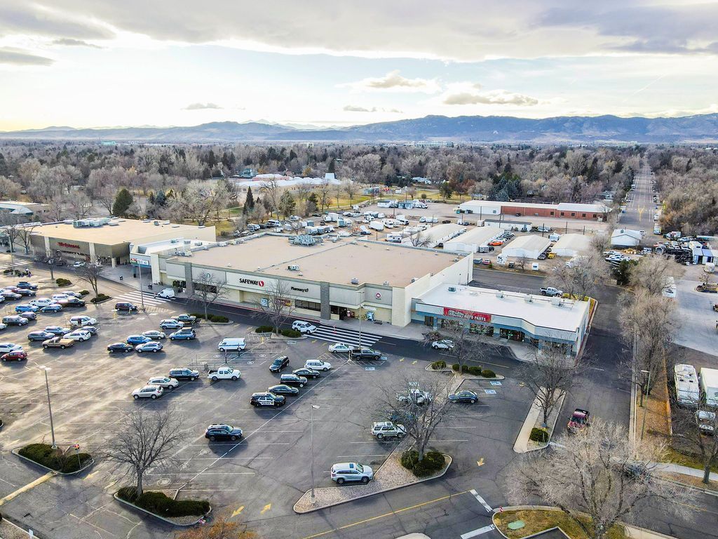 Aerial view of a shopping center with cars in the parking lot and trees in the background under a cloudy sky.