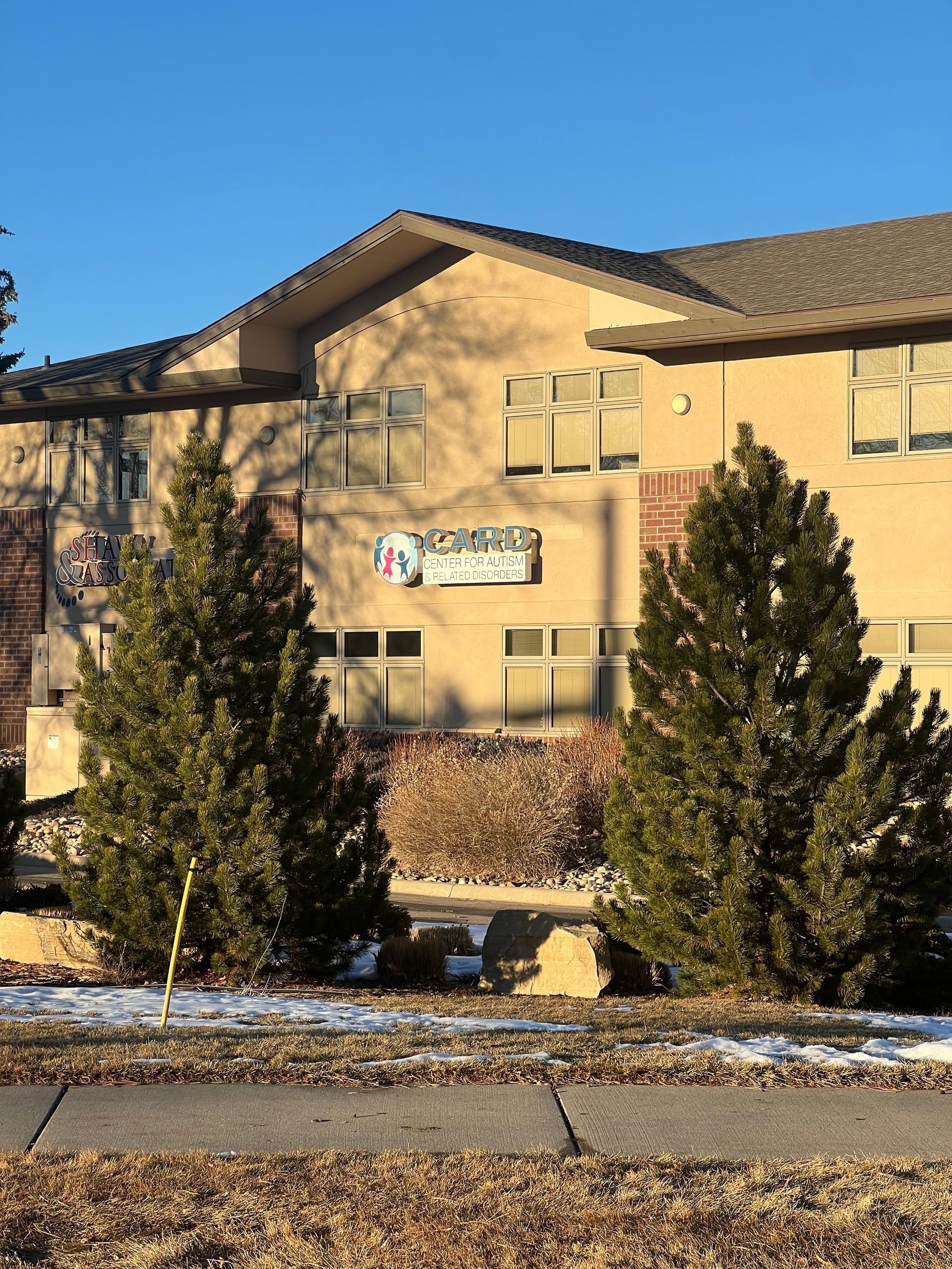 Two-story tan building with dark roof, evergreen trees in front, and a sign. Blue sky.