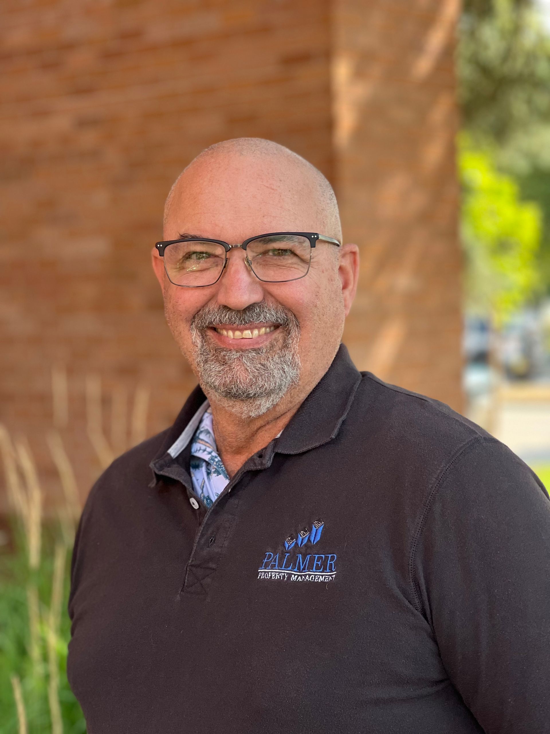 Man with glasses smiles, wearing a black shirt, outdoors near brick wall and greenery.