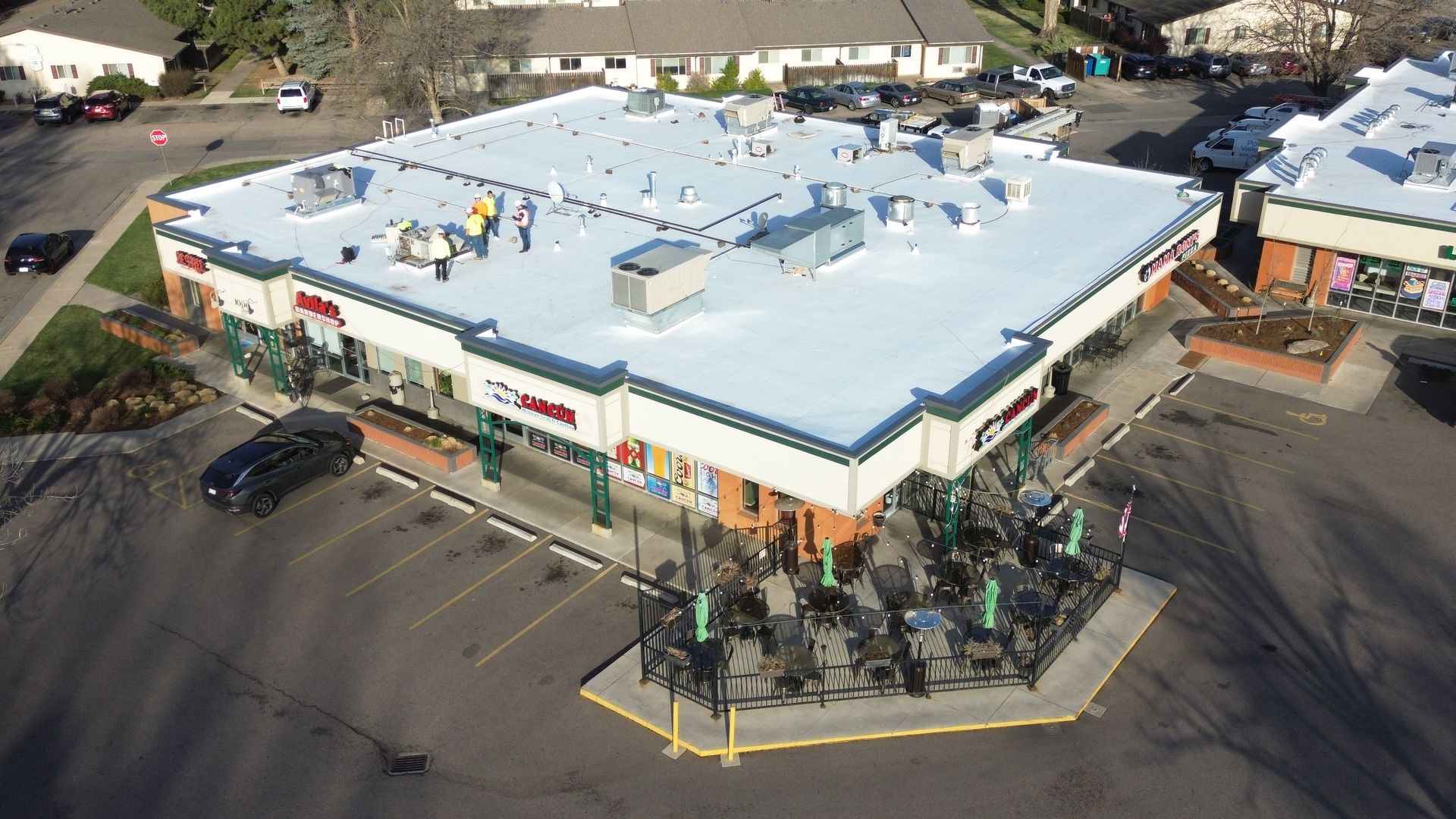 Aerial view of a commercial building with outdoor seating; surrounded by trees and a parking lot. Dusk sky.