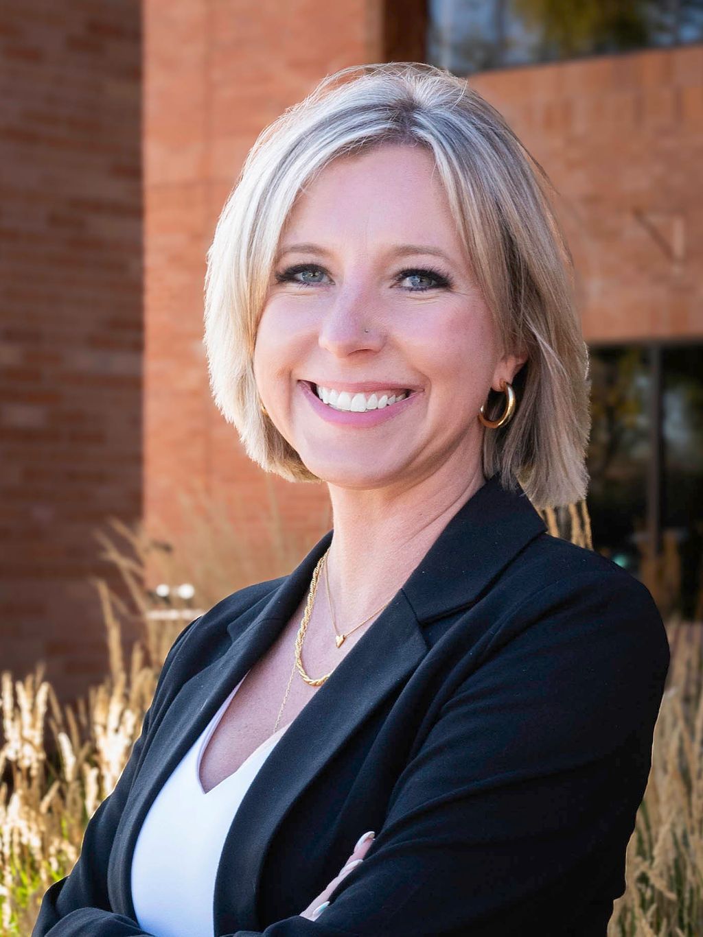 Woman with blonde hair smiles, wearing a black blazer and white top, arms crossed. Standing outside a building.