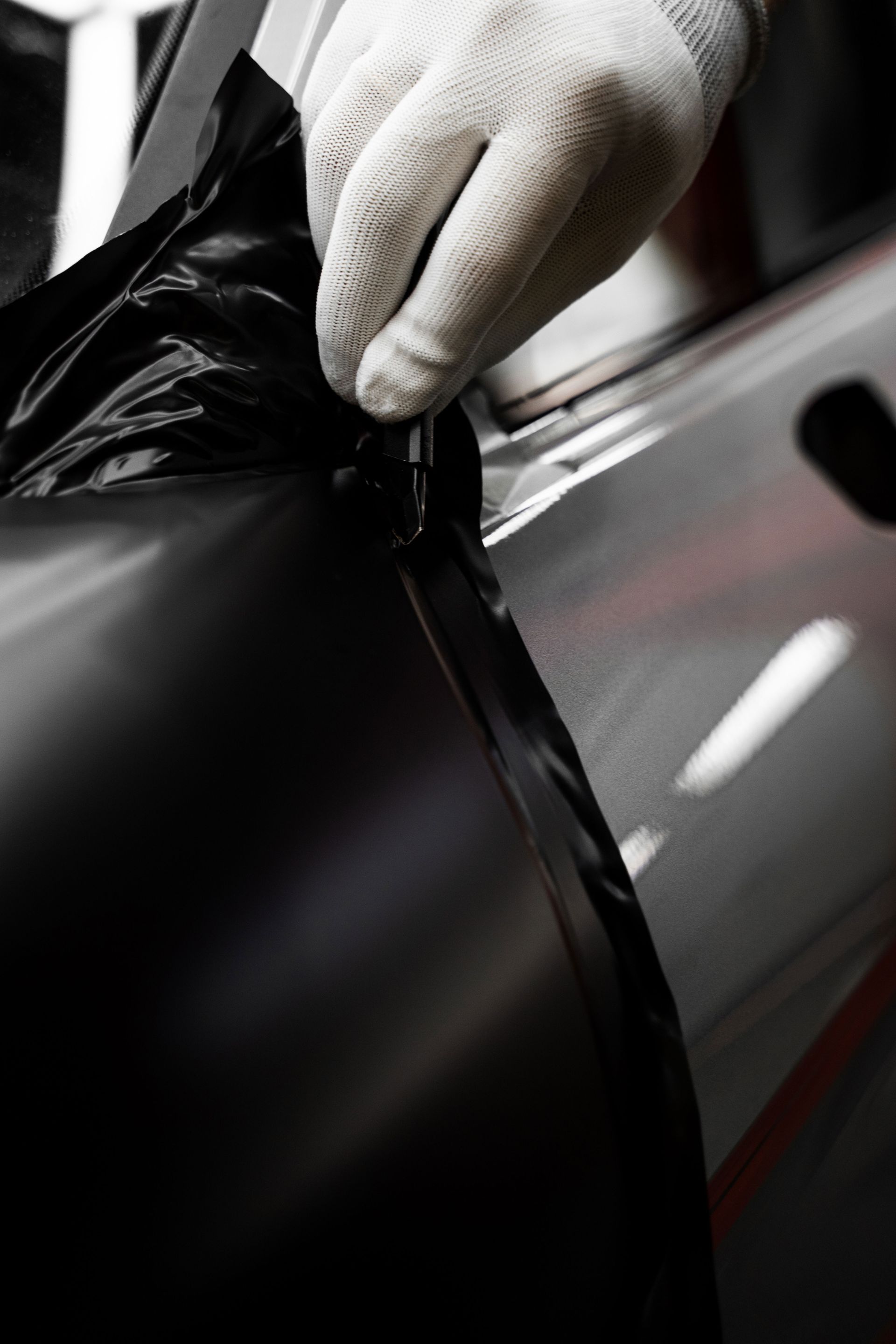 A gloved hand uses a tool to apply a black, matte vinyl wrap to the door of a silver vehicle.