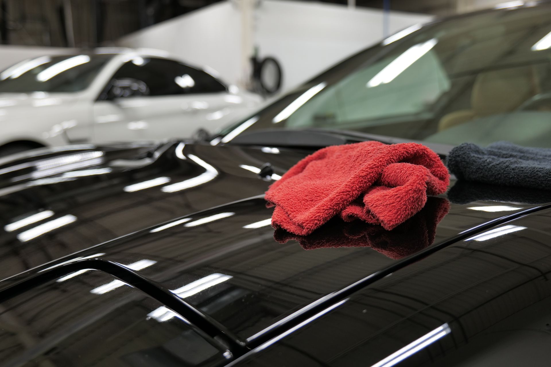 A red microfiber towel rests on the reflective, polished hood of a black car in a garage.