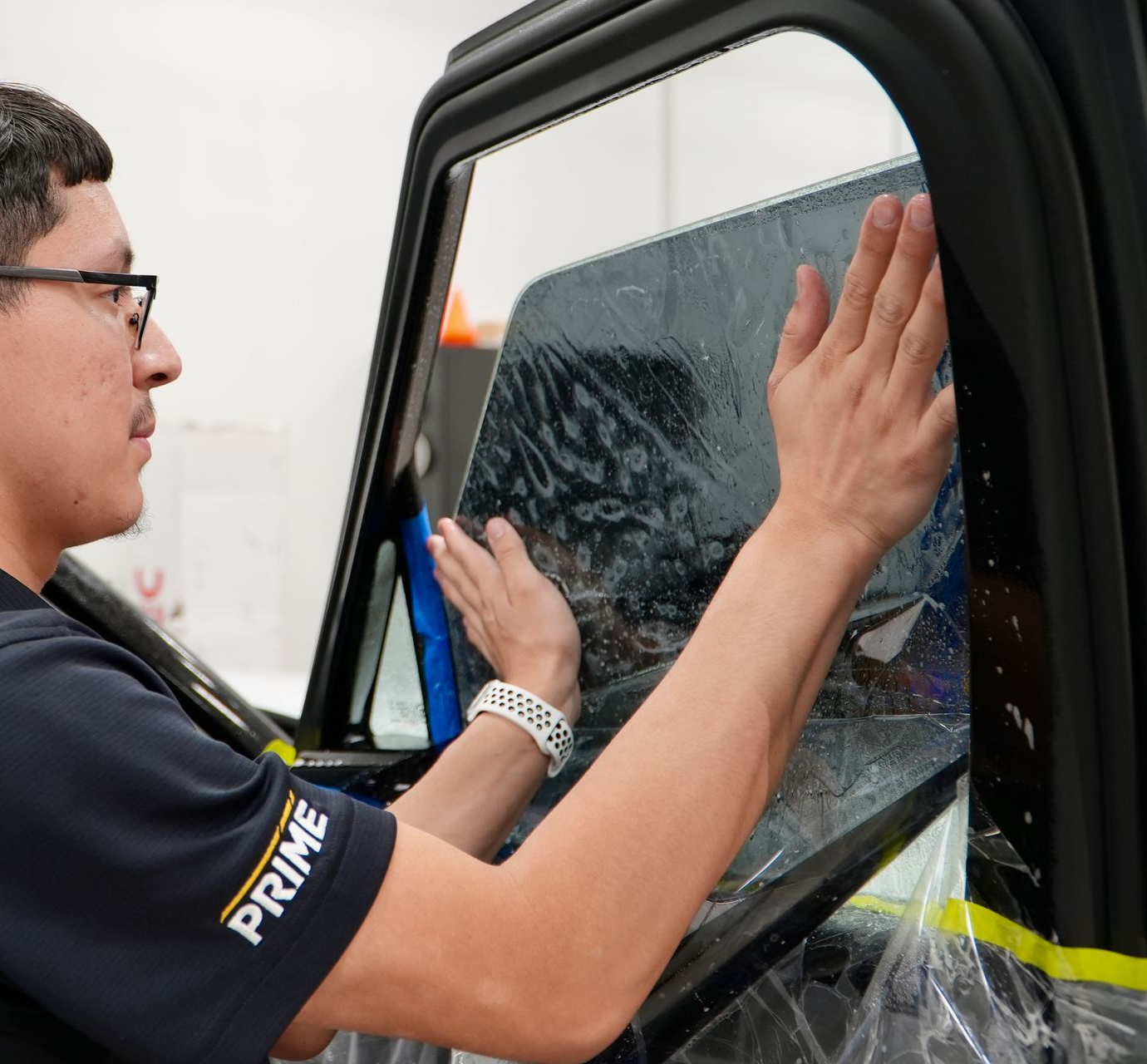 A hand wearing a black glove cleans a dark blue car hood using a red and white microfiber cloth and soapy foam.