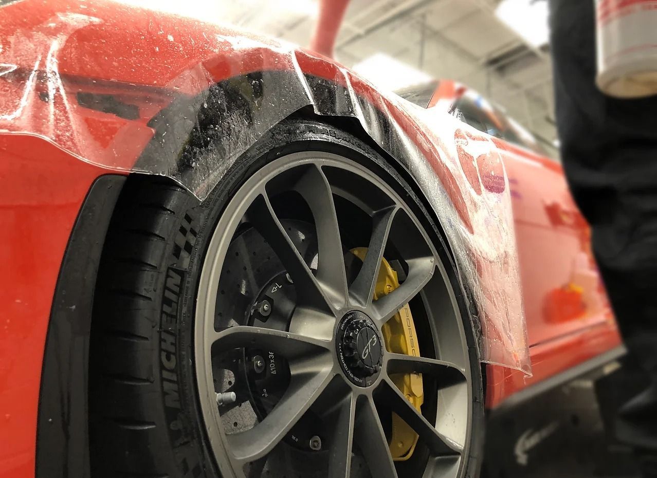 A close-up view of a clear protective film being applied to the red fender of a vehicle with a black wheel and yellow caliper.