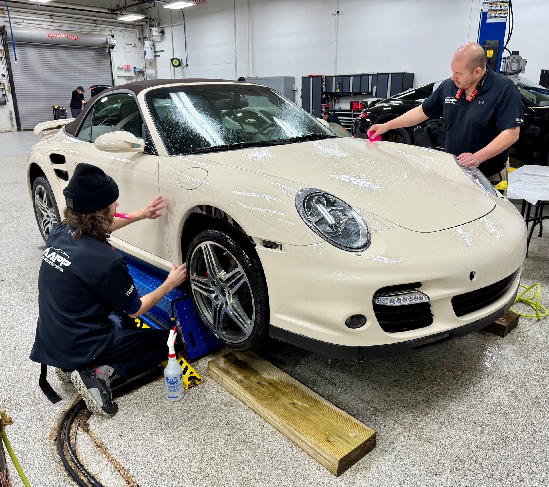 Two people in a garage working on a cream-colored Porsche convertible, applying a protective film to its exterior.
