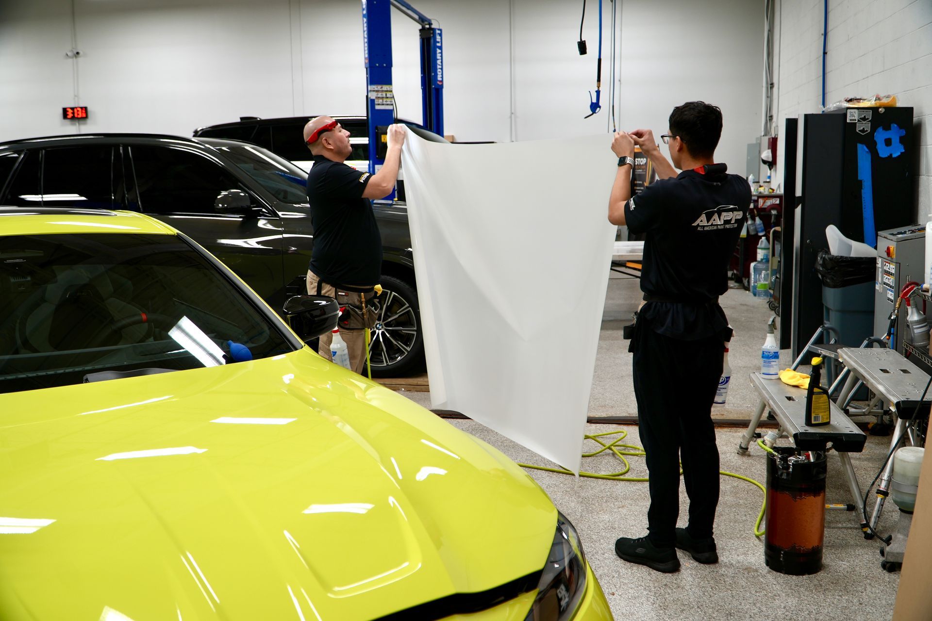 Two workers in a repair shop hold up a large sheet of white film in front of a bright yellow car.