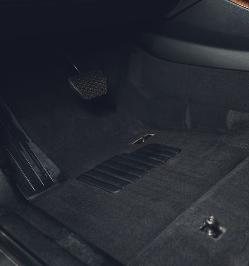 The driver-side car interior floor, showing dark gray carpet, a brake pedal, and a small, detached wire connector.
