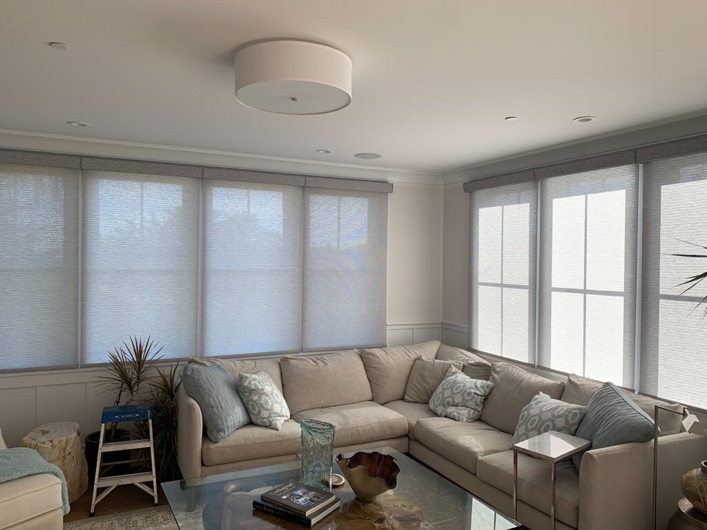 Bright living room with gray sectional sofa, large windows, and sheer blinds under a white ceiling light