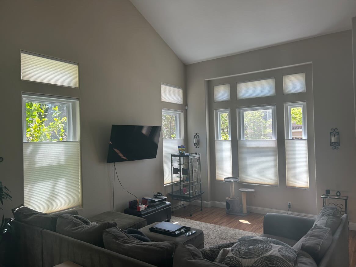 A high-ceilinged living room with a large corner sofa, a wall-mounted TV, and bright windows with pleated shades.