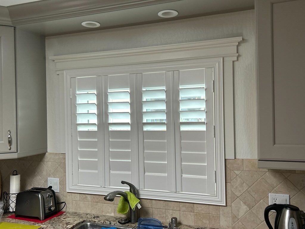 White louvered plantation shutters installed on a kitchen window with decorative trim, set above a sink and countertop.