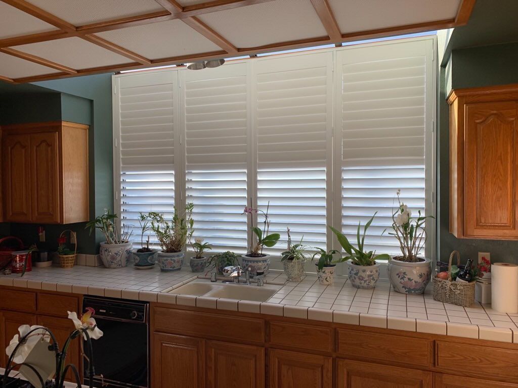 A kitchen sink area features tiled counters, oak cabinets, and white shutters covering a large window with potted plants.