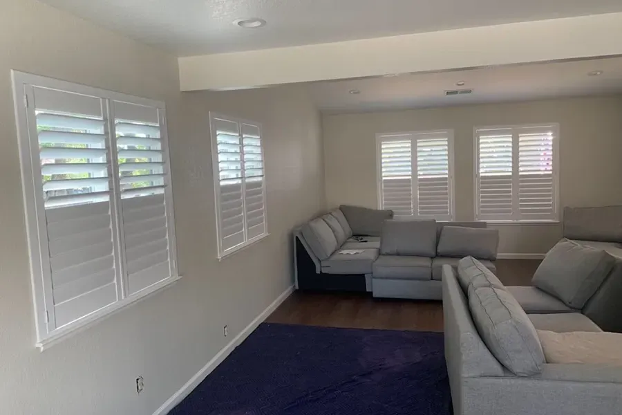 Bright living room with gray sectional sofas, white plantation shutters, and a dark blue rug.