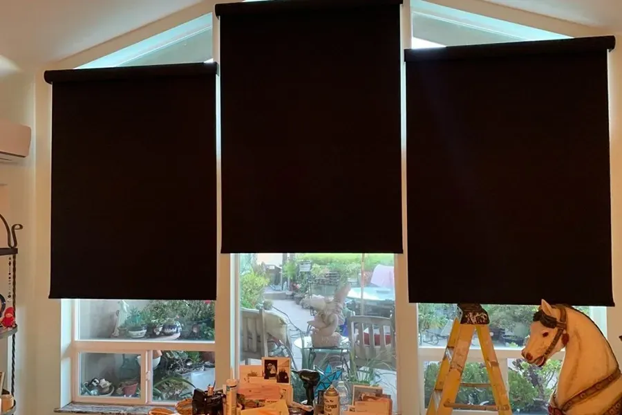 Three dark window shades cover a bright room, with a window view and a step ladder below.