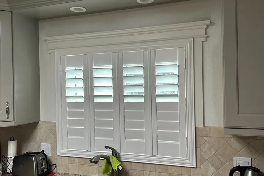 Kitchen window with white plantation shutters above a countertop and tile backsplash.