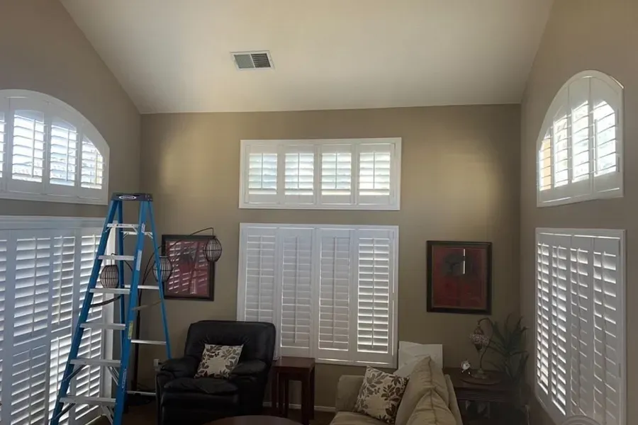 Bright living room with white plantation shutters, a blue ladder, black chair, and beige sofa against cream walls