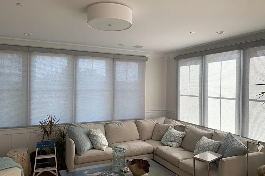 Bright living room with a beige sectional sofa, large windows with blinds, and a round ceiling light.