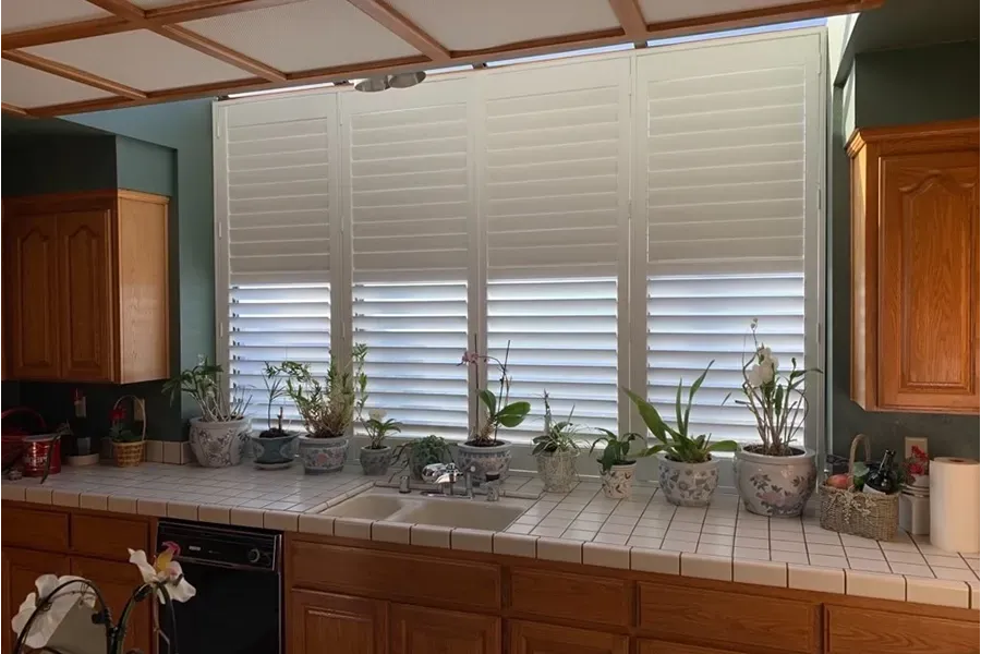Kitchen window with white shutters, tiled counter, and potted plants in front of wooden cabinets