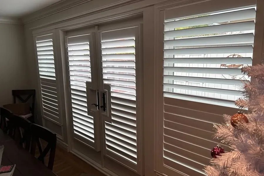 Sunlit living room windows with white plantation shutters and a dining table on the left