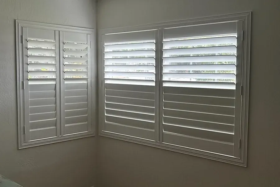 White plantation shutters on two corner windows in a beige room, partially open to daylight