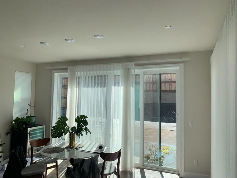 A dining area with a glass table, chairs, a potted Monstera plant, and sheer white curtains over a sliding glass door.
