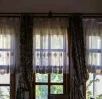 A bay window with three sets of white horizontal blinds partially drawn, showing a view of a street and cars outside.