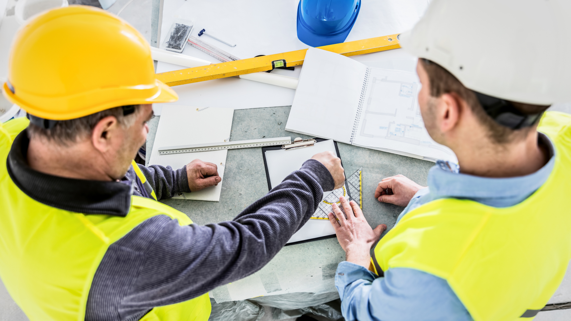 Two construction workers in yellow vests and hard hats reviewing blueprints.