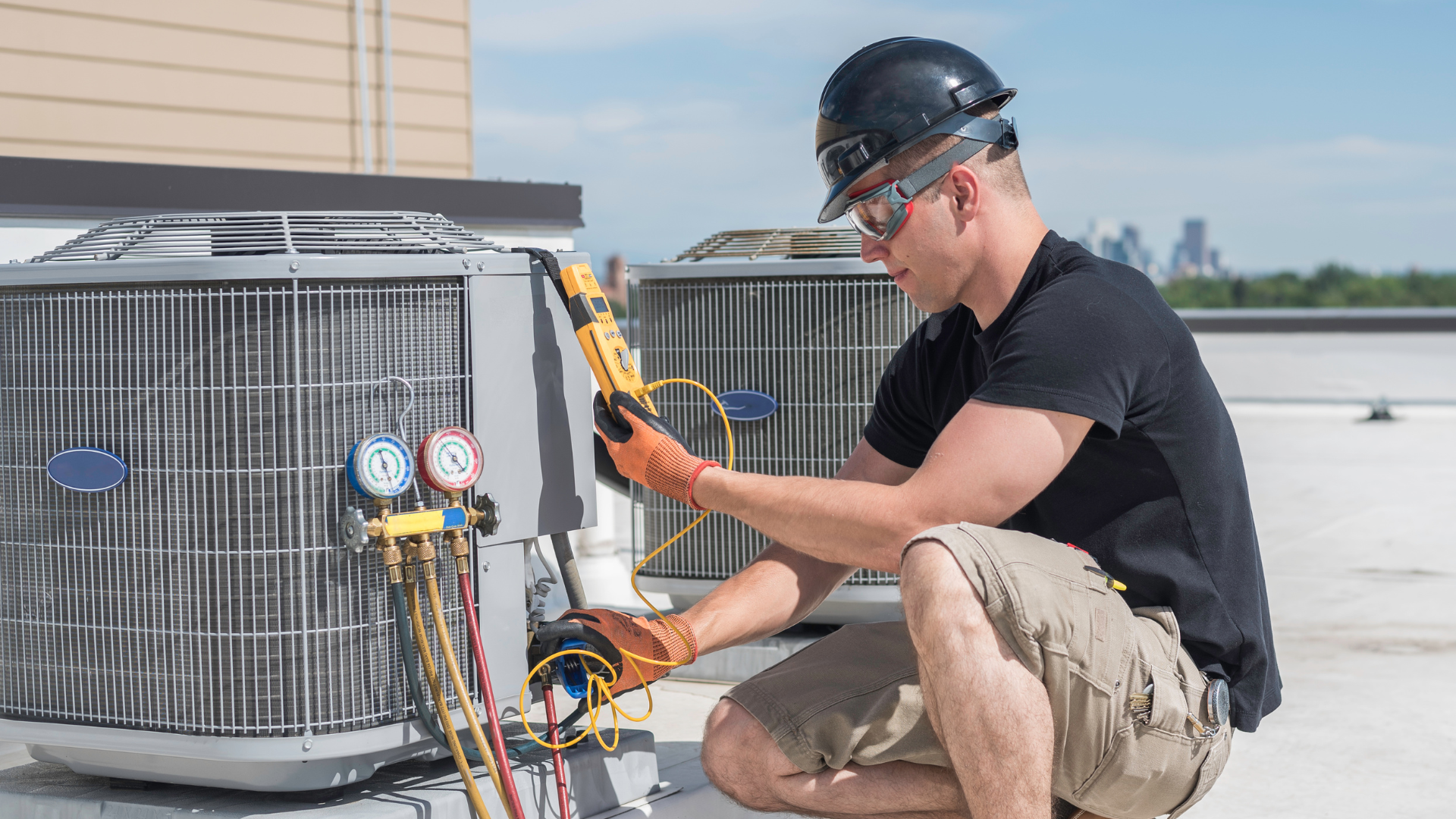 HVAC technician kneeling, inspecting an air conditioning unit on a rooftop, wearing safety gear.
