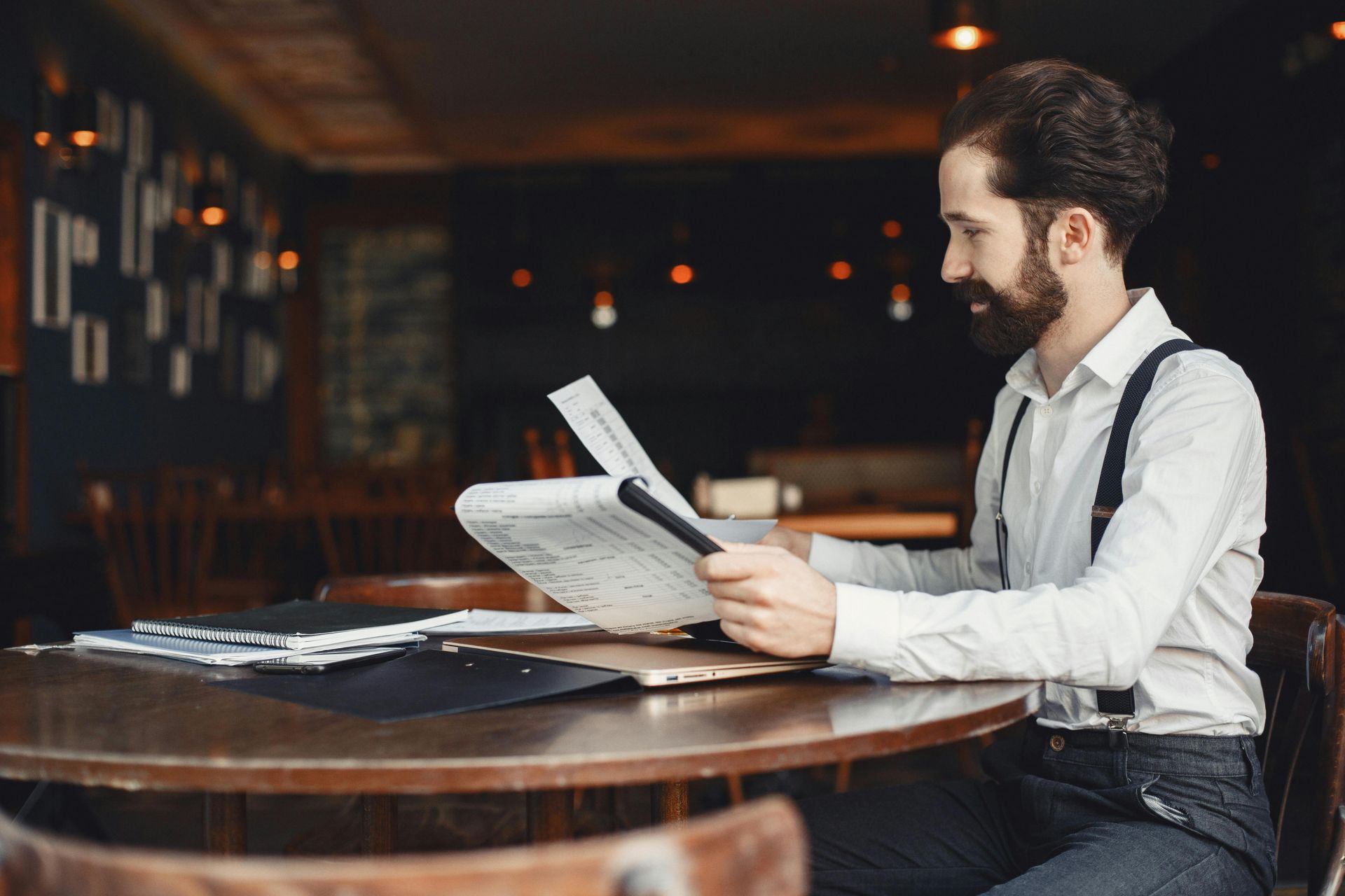 Man in white shirt and suspenders reading a newspaper at a table in a dim cafe