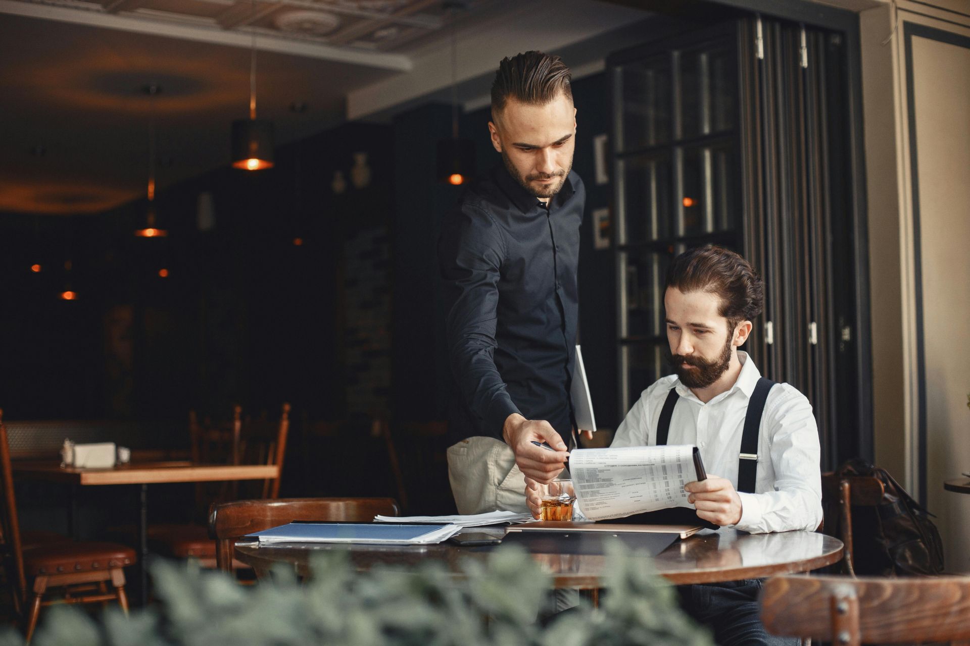 Two people reviewing a document at a restaurant table, one standing and pointing while the other sits and reads.