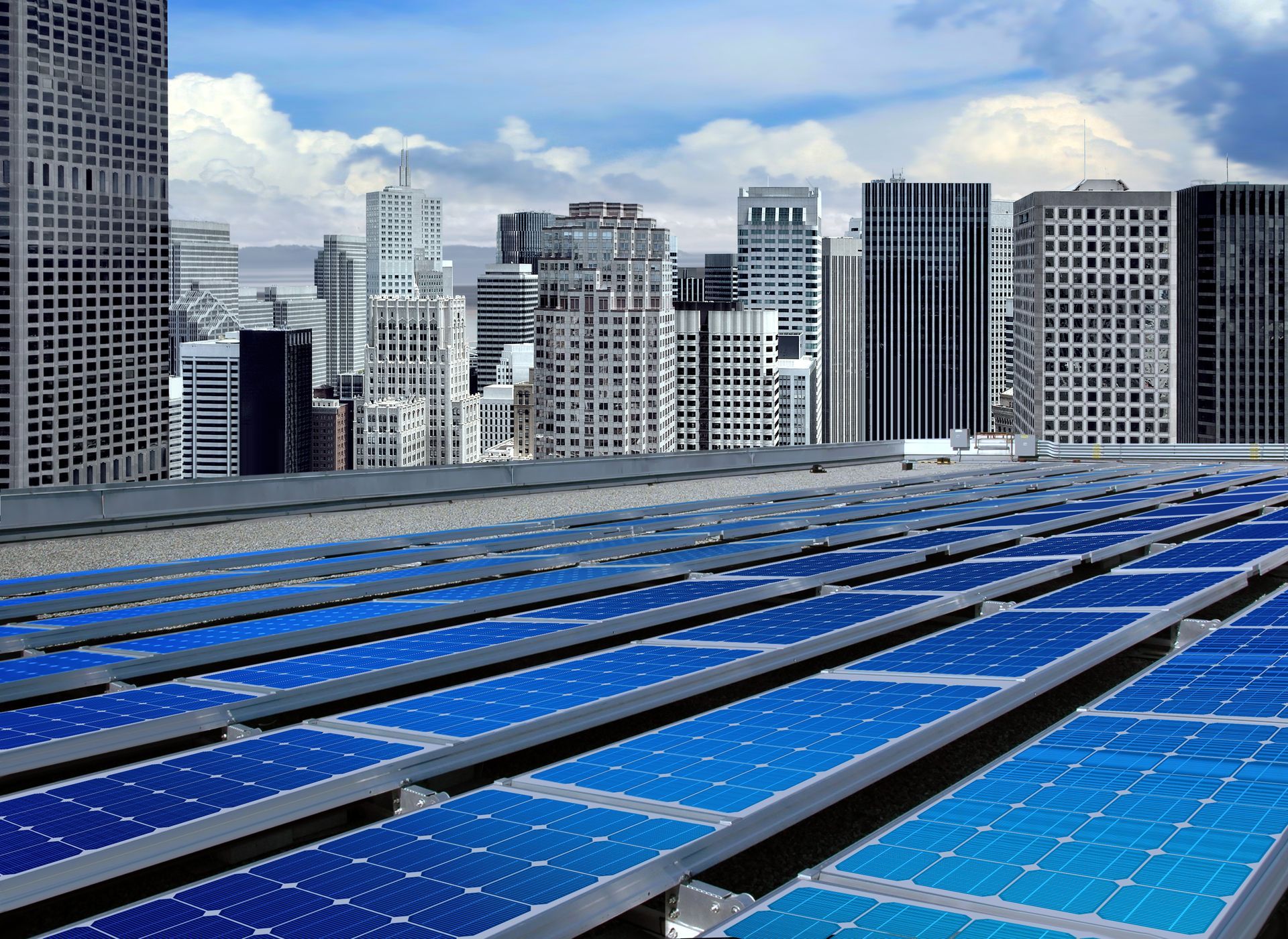 Solar panels on a building rooftop with a city skyline in the background. Blue panels against a blue sky.
