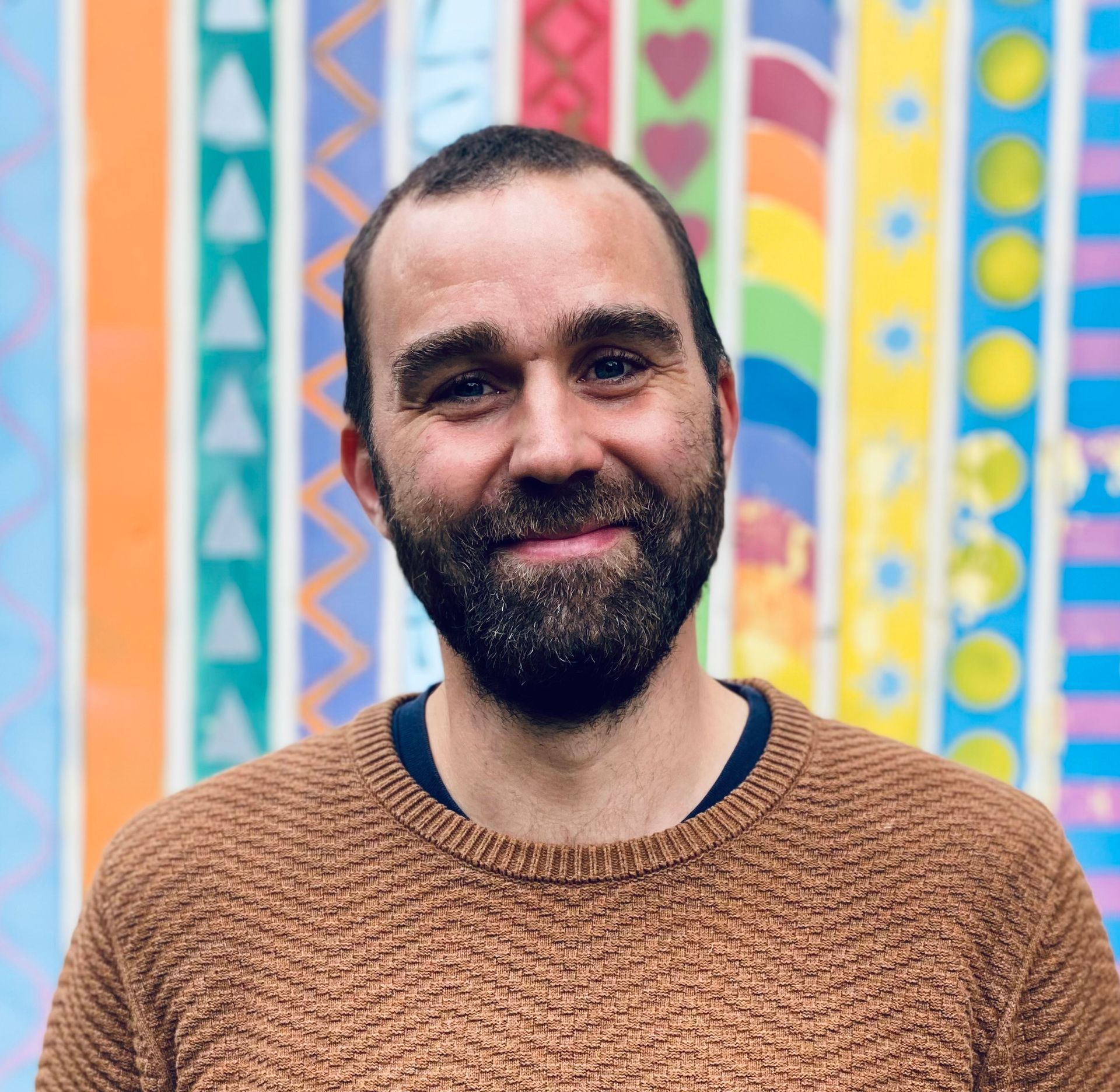 Ein lächelnder Mann in einem braunen Pullover steht vor einem farbenfrohen geometrischen Wandbild.