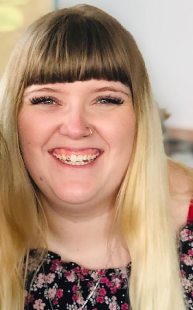 Lächelnde Person mit blonden Haaren und Pony, die ein geblümtes Oberteil trägt, vor einem weich verschwommenen Hintergrund