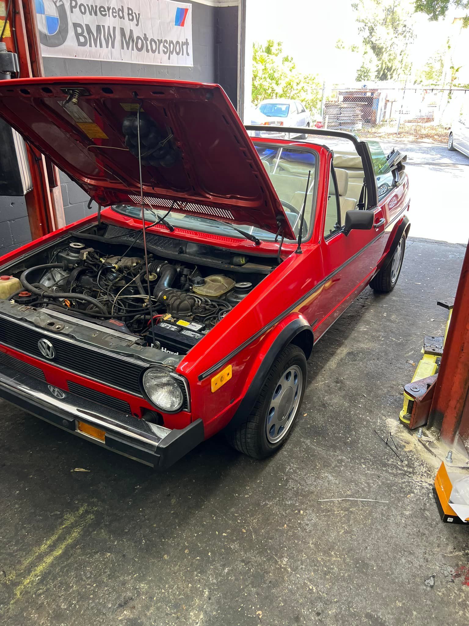 Red vintage car with hood open, showing engine bay, parked in a garage or workshop.