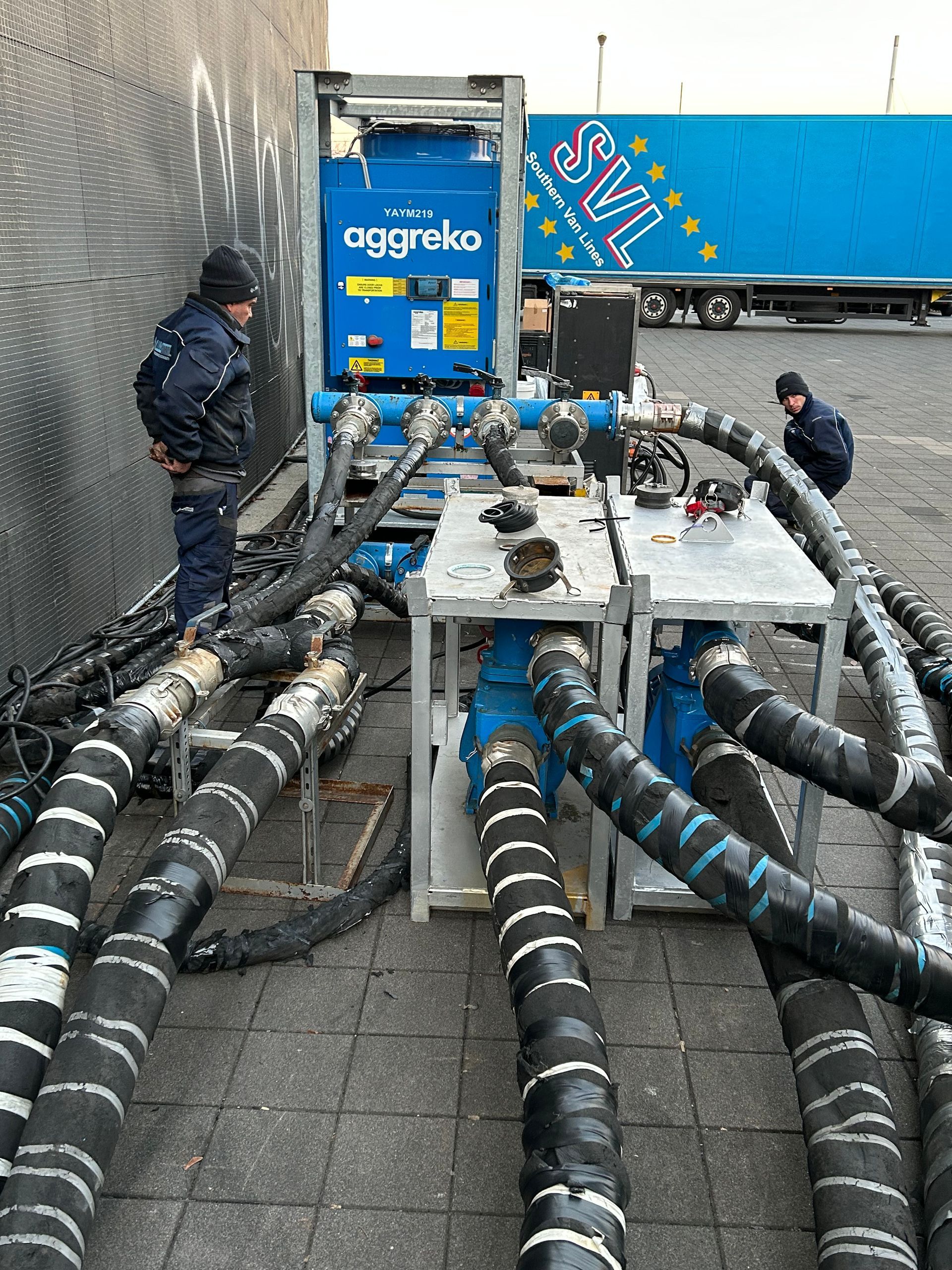 Dos hombres están trabajando en una máquina con un camión azul al fondo.