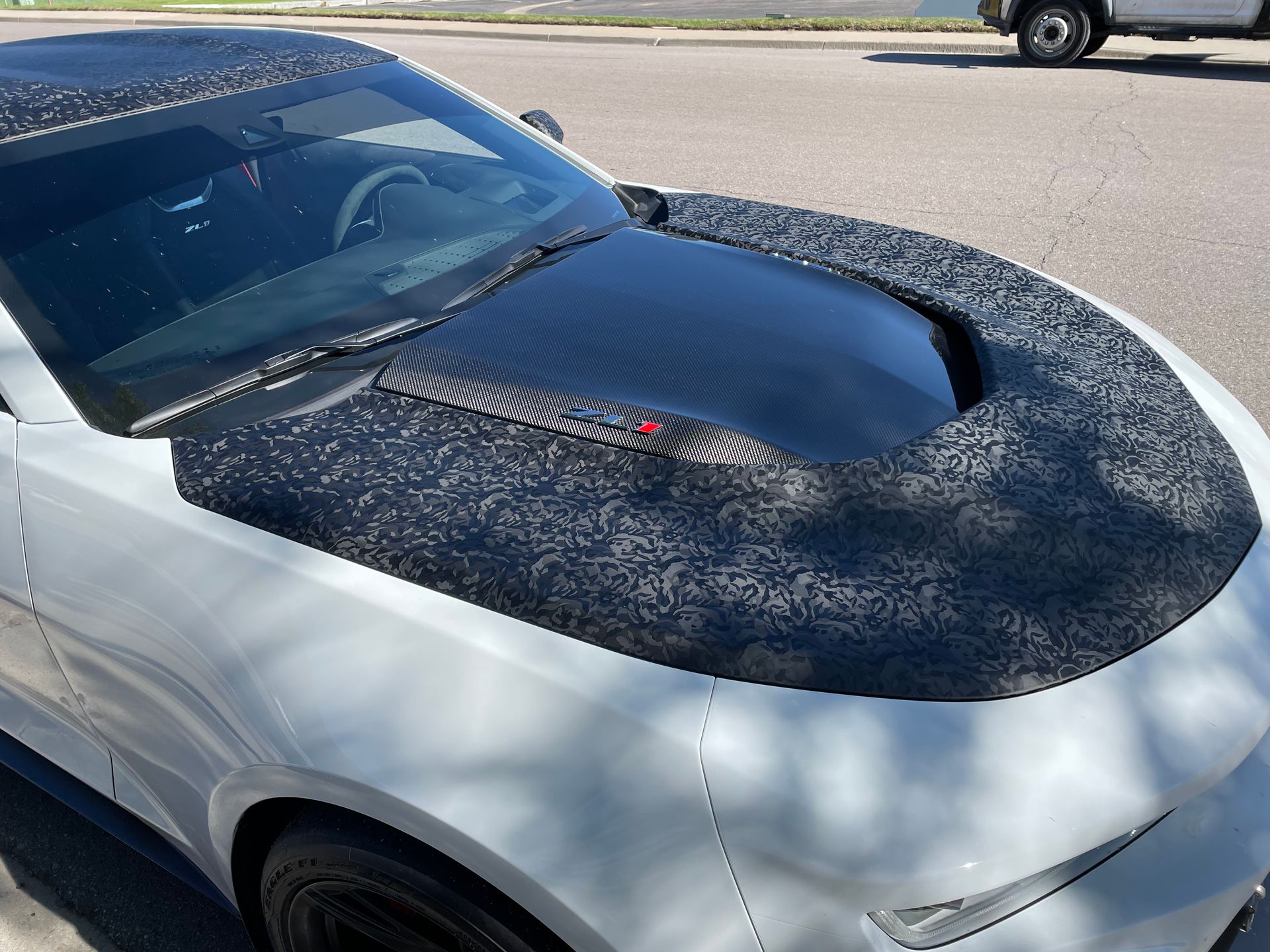 Silver car with a black hood covered in dark speckled wrap or debris, parked outdoors in sunlight.