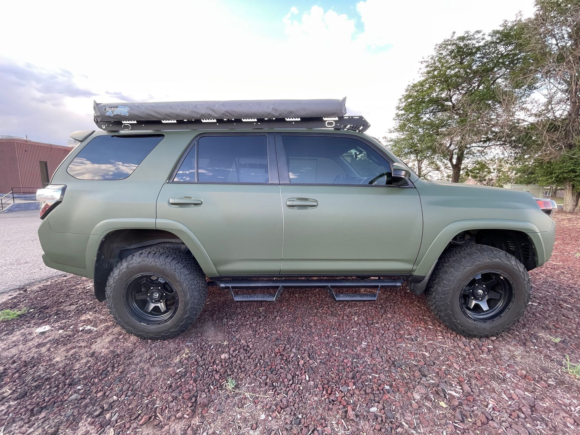 Olive green off-road SUV with roof rack parked on gravel under trees at sunset